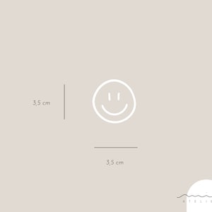 May include: A white smiley face icon on a light gray background. The icon is 3.5 inches wide and 3.5 inches tall. The text "atelier" is written in black below the icon.