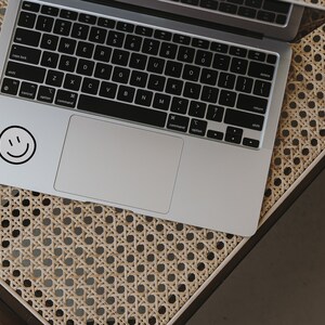 May include: A silver laptop computer with a black keyboard and a trackpad. A black and white smiley face sticker is on the top left corner of the laptop.