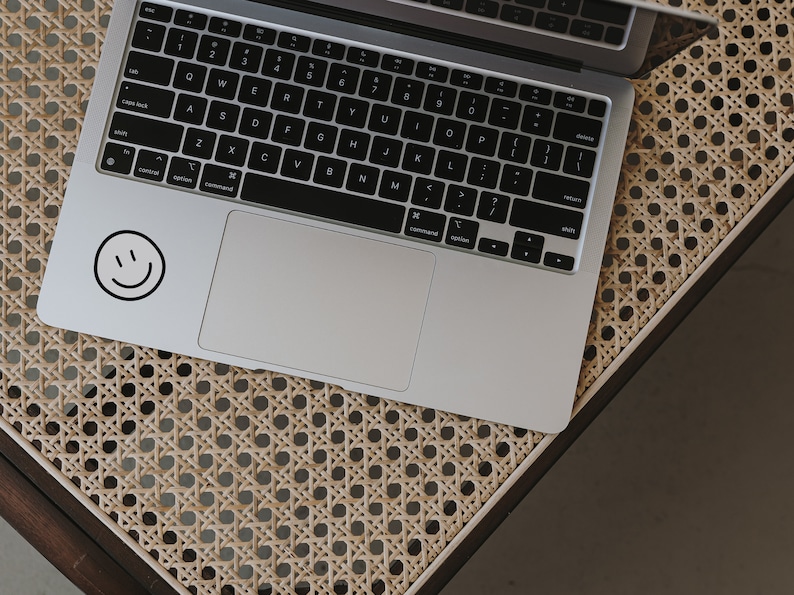 May include: A silver laptop computer with a black keyboard and a trackpad. A black and white smiley face sticker is on the top left corner of the laptop.