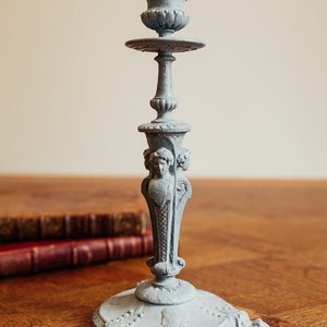 Può includere: Un candelabro in metallo grigio con un design elaborato. Il candelabro ha una base rotonda con tre piedi e uno stelo alto e sottile con una parte superiore decorativa. Lo stelo presenta un design scolpito di una testa di donna.