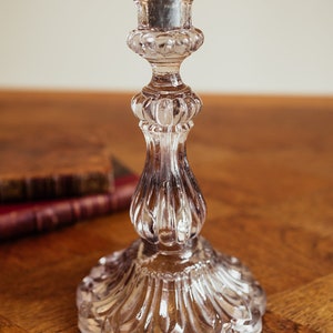 Può includere: Un candelabro di vetro trasparente con una candela grigia. Il candelabro ha un design dettagliato e ornato con una base ampia e un fusto scanalato. Il portacandele è su una superficie di legno.