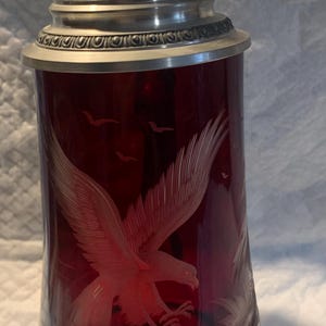 May include: A ruby red glass stein with a detailed metal lid and base. The body of the stein features an etched design of an eagle in flight. The text "Handarbeit Made W. Germany" is visible on a small label.
