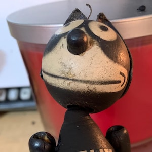 May include: A black wooden Felix the Cat figurine with a white face and black eyes. The figurine is standing with its arms raised and has a tail that is segmented. The figurine has the word "FELIX" written in gold letters on its chest.