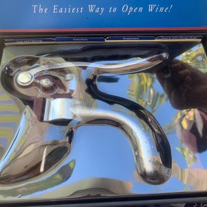May include: A chrome wine opener in a black box. The opener has a curved handle and a lever mechanism. The box has the text "The Easiest Way to Open Wine!" in white letters on a blue background.