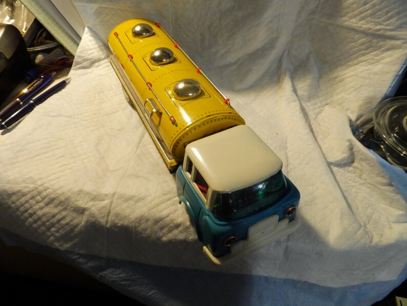 Pode incluir: Um caminh&atilde;o de brinquedo vintage com um tanque amarelo e uma cabine azul. O tanque tem tr&ecirc;s tampas prateadas no topo. O caminh&atilde;o &eacute; de metal e tem um telhado branco.