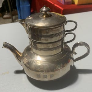 May include: Silver-plated coffee pot with a lid and spout. The pot has a round base and a cylindrical upper section with two handles. The lid is topped with a small, gold-colored knob. The initials "MMP" are engraved on the base.