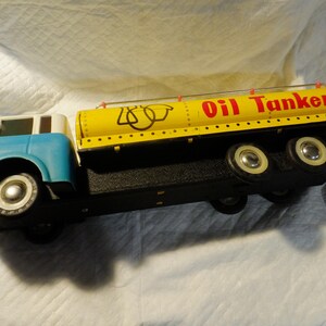 Pode incluir: Um caminh&atilde;o-tanque de brinquedo vintage com um tanque amarelo e uma cabine azul. O tanque tem um r&oacute;tulo vermelho "Oil Tanker" e marcas pretas. O caminh&atilde;o tem rodas brancas.