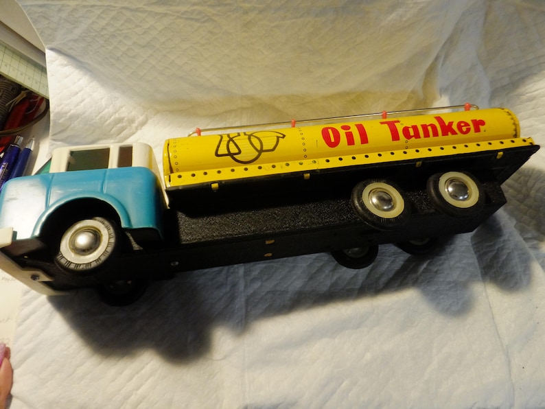 Pode incluir: Um caminh&atilde;o-tanque de brinquedo vintage com um tanque amarelo e uma cabine azul. O tanque tem um r&oacute;tulo vermelho "Oil Tanker" e marcas pretas. O caminh&atilde;o tem rodas brancas.