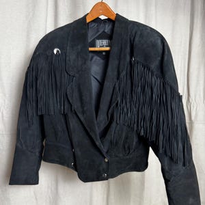 May include: Black suede jacket with fringe detailing. The jacket features a notched collar, button closures, and decorative silver-tone accents. The label reads "OUTERBOUND".