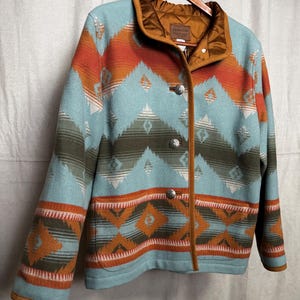 May include: A turquoise, orange, and olive green patterned jacket with silver buttons and a brown collar. The jacket is hanging on a wooden hanger. The jacket has a geometric design.