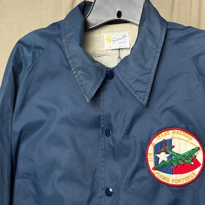 May include: A navy blue nylon jacket with a button-up front and a pointed collar. The jacket features a circular patch on the chest with the text "Texas Raiders Flying Fortress" and an image of a green airplane. The jacket has a "Swingster Jackets" label.