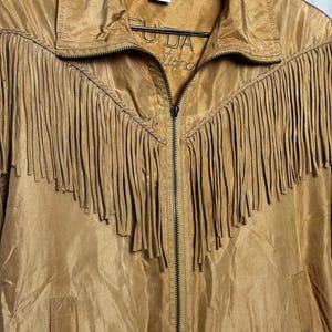 May include: A tan jacket with a zipper closure and fringe detailing across the chest. The jacket has a collared neckline and a label with text. The material has a slight sheen.