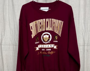 May include: A maroon sweatshirt with the words "SOUTHERN CALIFORNIA" in gold lettering. Below the text is a crest with the word "TROJANS" and the text "EST. 1880 Los Angeles, Calif". The sweatshirt is hanging on a metal hanger.