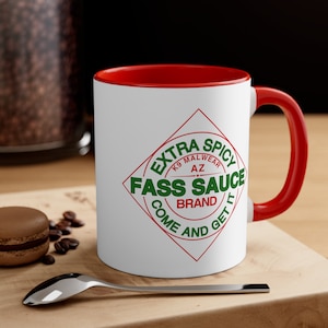 May include: White ceramic mug with a red rim and handle. The mug features a red diamond with the text "EXTRA SPICY K9 MALWEAR AZ FASS SAUCE BRAND COME AND GET IT" in green letters.