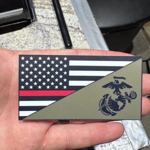 May include: A rectangular patch featuring a split design. The top left half displays a black and white American flag with a thin horizontal red stripe. The bottom right half is olive green and features a dark blue United States Marine Corps emblem.