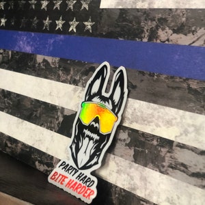 K&ouml;nnte beinhalten: Ein schwarz-wei&szlig;er Aufkleber eines Hundes, der eine Sonnenbrille mit Regenbogen-Schimmer tr&auml;gt. Die Zunge des Hundes h&auml;ngt heraus. Der Aufkleber tr&auml;gt den Text "Party Hard Bite Harder". Der Aufkleber befindet sich auf einem Hintergrund einer abgenutzten amerikanischen Flagge mit einem blauen Streifen.