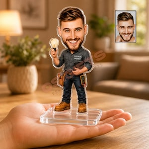 May include: A custom acrylic bobblehead figurine of an electrical engineer with a beard, wearing a dark gray shirt with the text 'ELECTRICAL ENGINEER', blue jeans, and brown boots, holding a light bulb and tools on a clear square base.