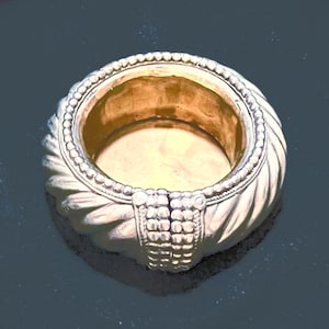 May include: A gold-colored, circular ring dish with a textured, braided design around the outer edge. The dish has a detailed, rectangular pattern at the base and a smooth, golden interior.