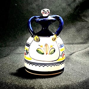 May include: A ceramic bell shaped like a woman with a blue dress, yellow skirt, and green and orange floral design.