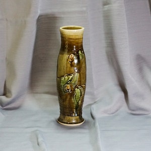 May include: A brown ceramic vase with a green and yellow floral design. The vase has a cylindrical shape with a flared lip and a rounded base.