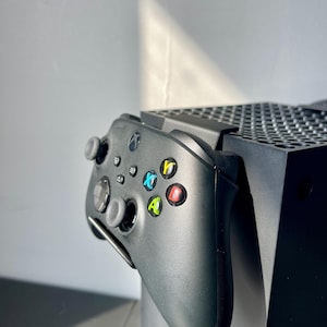 May include: A black Xbox Series X console and controller. The controller features coloured buttons and a directional pad. The console is positioned vertically, with the controller attached to the side.