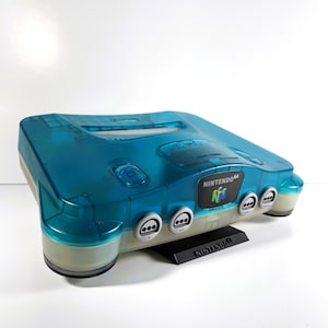May include: A translucent blue Nintendo 64 console with a black stand. The console has a clear plastic case and a grey Nintendo 64 logo on the front.