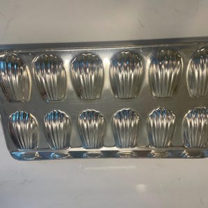 May include: A silver madeleine tin with 12 shell-shaped moulds. The rectangular tin has rounded edges and is made of reflective metal. The moulds have a fluted design.