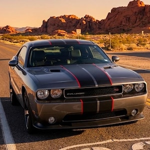 May include: A gray Dodge Challenger with black and red racing stripes parked on a road marked with a white Route 66 shield. In the background, a vintage Hotel Motel sign stands near red rock desert formations under a clear sunset sky.
