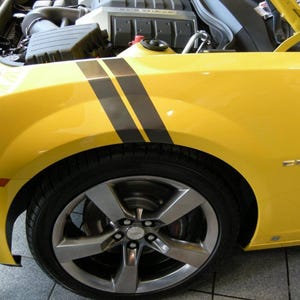 Puede incluir: Un Chevrolet Camaro amarillo con rayas de carreras negras y una llanta de aleación negra y plateada. El capó está abierto, revelando el motor.