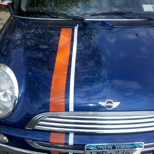 Puede incluir: Un Mini Cooper azul con franjas de carreras naranjas y blancas en el capó. El coche tiene detalles cromados, una parrilla delantera y una matrícula de Nueva York. Los espejos laterales tienen un diseño de la Union Jack.