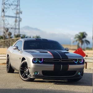May include: A silver Dodge Challenger coupe featuring dual black racing stripes with thin red accents running down the center of the hood. The vehicle is equipped with circular headlights and fog lights that emit a vibrant, multi-colored LED glow.