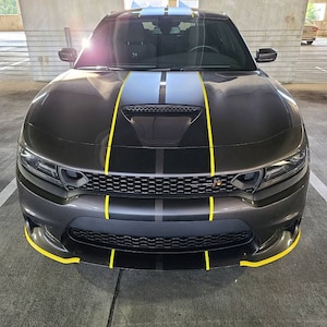 May include: A black Dodge Charger with yellow and grey racing stripes on the bonnet. The car has a black grille and black headlights.
