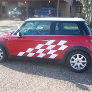 Puede incluir: Un Mini Cooper rojo con una calcomanía de raya de carreras a cuadros blancos. El coche tiene un techo blanco y ruedas negras.