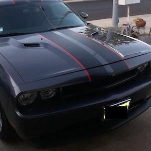 May include: A dark gray muscle car featuring a matte black center hood stripe flanked by thin red pinstripes. The vehicle has a prominent front grille with circular headlights, a vented hood, and a front license plate holder with a blacked-out plate.