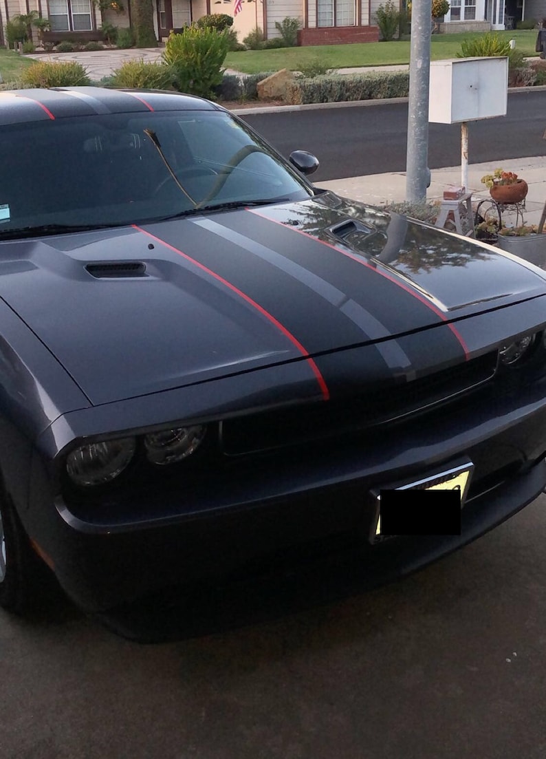 May include: A dark gray muscle car featuring a matte black center hood stripe flanked by thin red pinstripes. The vehicle has a prominent front grille with circular headlights, a vented hood, and a front license plate holder with a blacked-out plate.