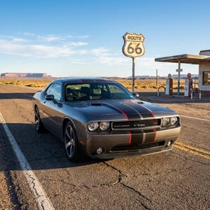 May include: A dark gray Dodge Challenger coupe featuring dual black racing stripes with thin red accents running down the hood and roof. The vehicle is parked on an asphalt road near a Route 66 sign and a vintage gas station in a desert landscape.