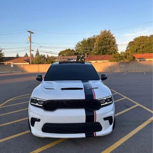 Puede incluir: Un SUV blanco con rayas de carreras negras y rojas en el centro. El vehículo tiene una parrilla delantera negra y detalles en negro. El coche está estacionado en una superficie pavimentada con líneas amarillas de estacionamiento, bajo un cielo azul claro.