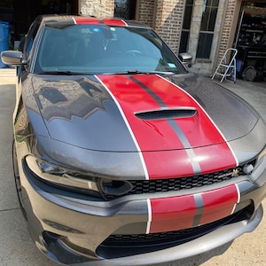 May include: A grey Dodge Charger with a black and red racing stripe down the bonnet. The car has a black grille and black headlights.