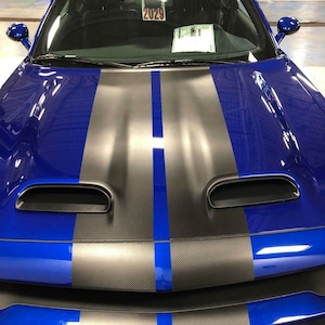 Può includere: Un'auto blu con strisce da corsa nere e blu. L'auto ha un cofano in fibra di carbonio nero e un paraurti anteriore nero. L'auto ha una griglia nera e uno spoiler nero. L'auto ha un parabrezza nero e un lunotto posteriore nero. L'auto ha un interno nero. L'auto ha una targa nera con il numero 2029.