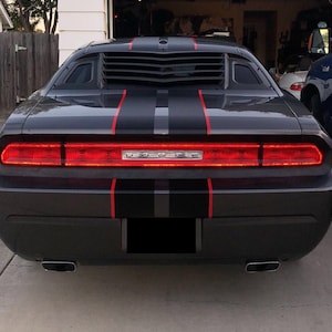 May include: A dark gray Dodge Challenger seen from the rear, featuring a full-width red LED taillight assembly with the Dodge logo in the center. The car has black racing stripes with thin red pinstripes and black rear window louvers.