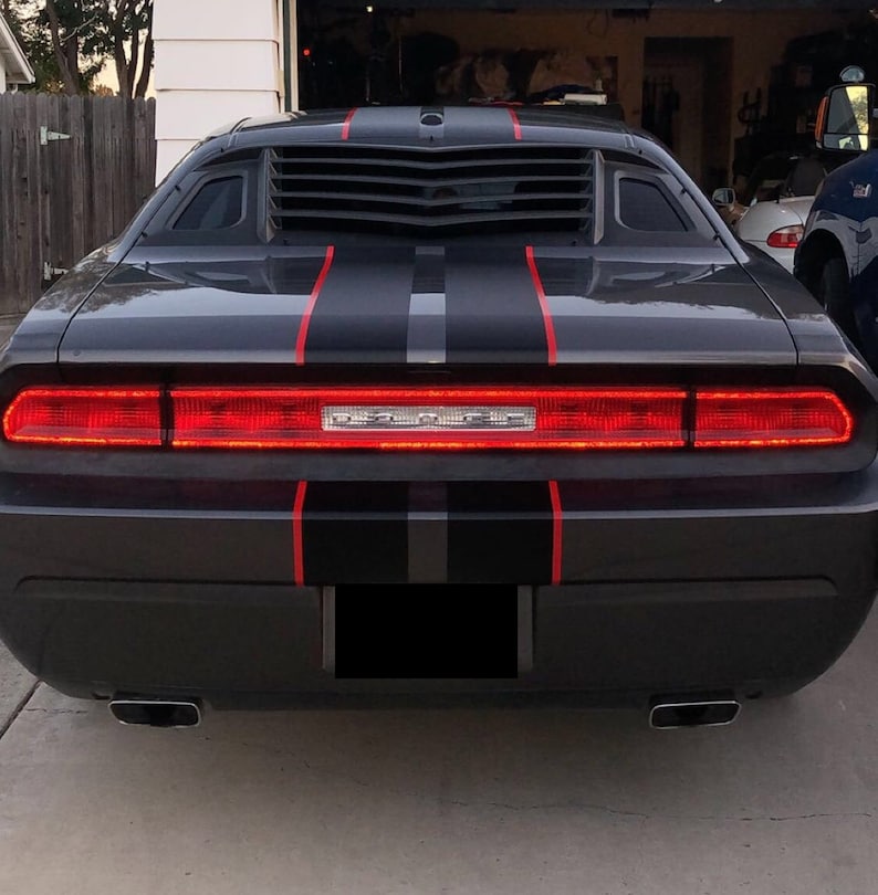 May include: A dark gray Dodge Challenger seen from the rear, featuring a full-width red LED taillight assembly with the Dodge logo in the center. The car has black racing stripes with thin red pinstripes and black rear window louvers.