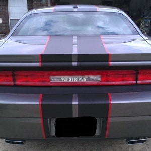 May include: The rear view of a gray car featuring a wide matte black racing stripe running down the center, accented by thin red pinstripes on both sides. The red taillight assembly spans the width of the trunk and displays the text A1 STRIPES.