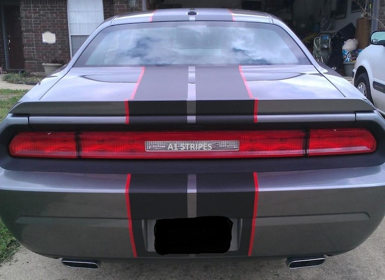 May include: The rear view of a gray car featuring a wide matte black racing stripe running down the center, accented by thin red pinstripes on both sides. The red taillight assembly spans the width of the trunk and displays the text A1 STRIPES.