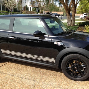 Puede incluir: Un Mini Cooper S negro con rayas de carreras plateadas y la palabra "COOPER S" en el lateral. El coche tiene ruedas negras y está aparcado en una entrada pavimentada.