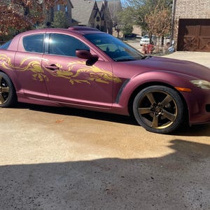 Puede incluir: Un Mazda RX-8 color burdeos con calcomanías de dragón doradas. El coche está estacionado en una entrada pavimentada.