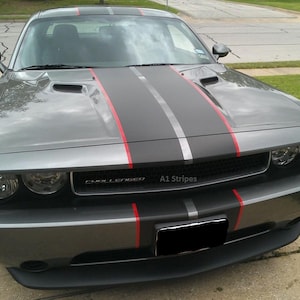 May include: A gray Dodge Challenger featuring a matte black center racing stripe flanked by thin red pinstripes running from the hood to the front bumper. The front grille displays the text Challenger and A1 Stripes with a black license plate.