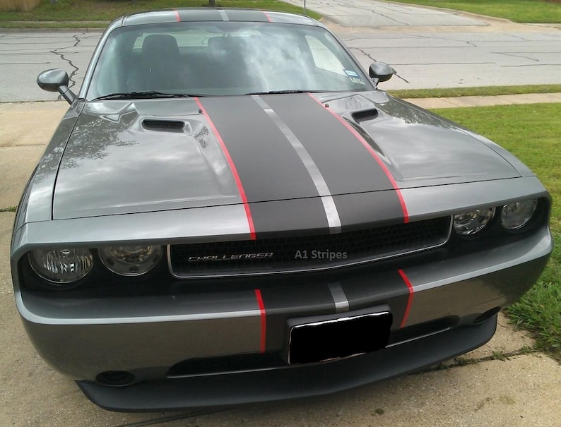 May include: A gray Dodge Challenger featuring a matte black center racing stripe flanked by thin red pinstripes running from the hood to the front bumper. The front grille displays the text Challenger and A1 Stripes with a black license plate.