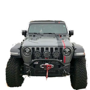 Puede incluir: Un Jeep Wrangler gris con un parachoques delantero negro y una franja roja y negra en el capó. El Jeep tiene un juego de luces todoterreno montadas en el parachoques delantero.