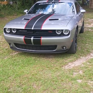 May include: A silver Dodge Challenger coupe featuring a black and red racing stripe running down the center of the hood and roof. The windshield displays the text Quicksilver. The vehicle has a black front grille with circular headlights.