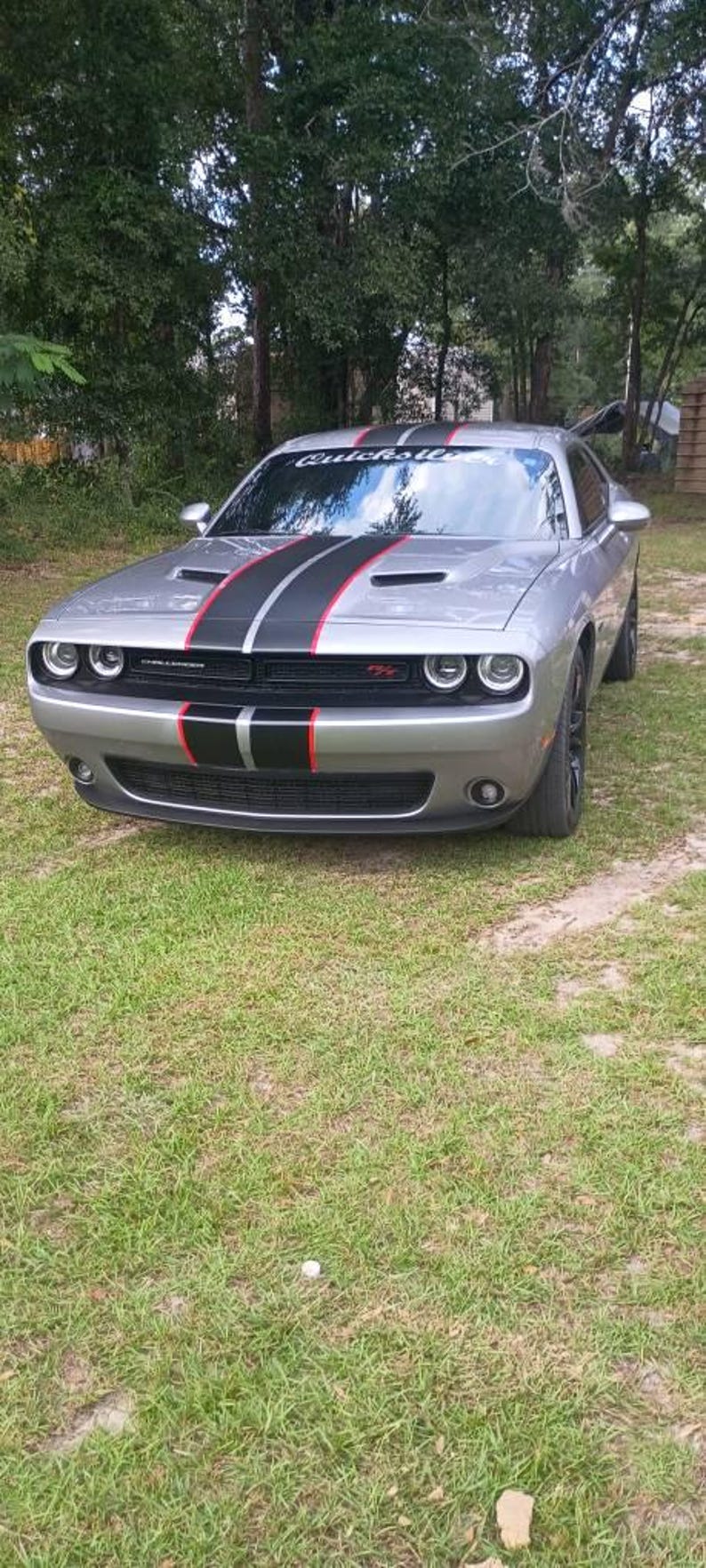 May include: A silver Dodge Challenger coupe featuring a black and red racing stripe running down the center of the hood and roof. The windshield displays the text Quicksilver. The vehicle has a black front grille with circular headlights.